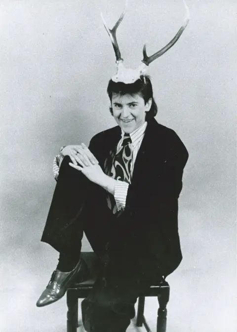 Handout/Embassy Theatre A black and white image of Joe Pasquale as a young man, wearing a black suit and comedy antlers. He is sitting on a stool and smiling with his hands on his raised right knee.