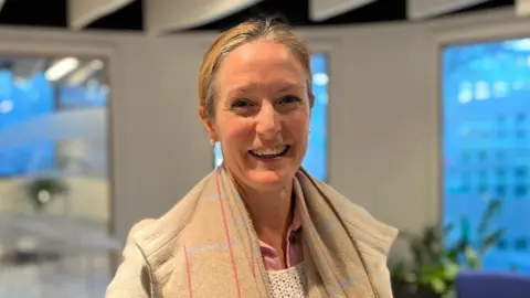 A smiling woman with her hair pulled back wearing a beige scarf and jacket