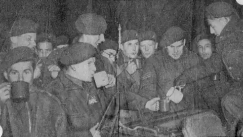 Danny Mason Follet de una fotografía granulada en blanco y negro de un periódico que muestra a los paracaidistas reunidos en un grupo bebiendo té. Están usando boinas. Muestra un momento justo antes de que entraran en aviones para ser dejados sobre líneas alemanas en el río Rhine en la Operación Varsity, marzo de 1945.