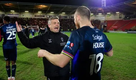 Chris Vaughan/Getty Images Two people stand on a football pitch after a match, facing each other with arms raised as they talk. One man is wearing a black padded jacket while the other wears a dark blue playing kit with the number 18 on the back. Additional players, red stadium seating and a crowd are in the background.