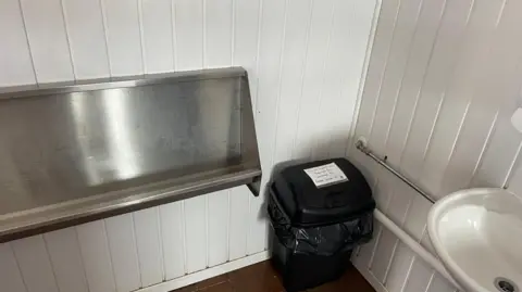 A picture of a small black bin with a piece of paper stuck to it in a corner. The walls are white. There is a sink to the right of the picture.