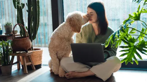 Getty Images Young woman kissing with her dog while using laptop, sitting on the floor, relaxing at home on a sunny afternoon.