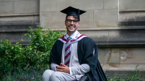 PA Media/University of Bristol Xander Van der Poll in his graduation gown and cap, sitting in his wheelchair smiling outside a university building