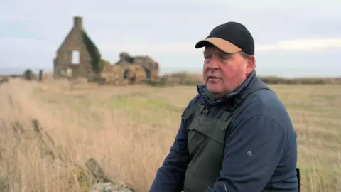 Alan Steven side on to the camera wearing a dark baseball cap with a brown peak, a blue fleece and waterproof dungarees. There is a dilapidated building in the field behind him.