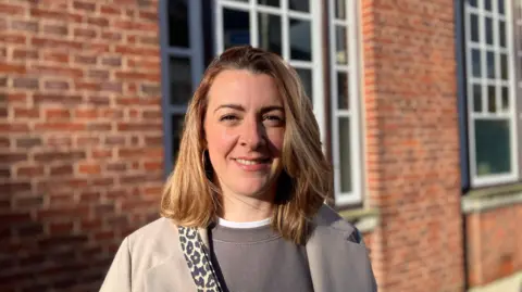 A woman with shoulder length blonde hair wearing a grey jumper and coat. 