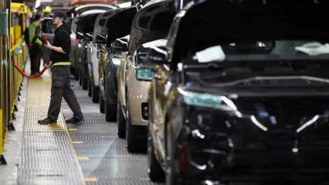 This generic image from 2022 shows a member of staff on the left working on the production line at Jaguar Land Rover's factory in Solihull. Cars are lined up with their bonnets up.
