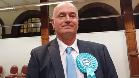 A middle-aged man wearing a light blue rosette and a suit stands in a public hall of some kind. His tie is also light blue.