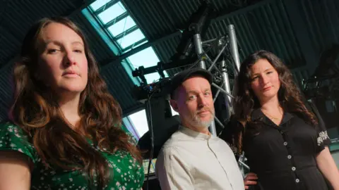 Ben Hughes Becky and Rachel Unthank stand either side of band member and producer Adrian McNally. The sisters have long, brown wavy hair with one wearing a green flowery top and the other a black button-up dress. McNally is wearing a dark flat-cap and light button-up shirt. He has a grey beard and moustache. They are standing in a warehouse-like building with a large skylight above and looking down into camera. Pictured behind them are some grey poles and other equipment which looks like a lighting rig. 