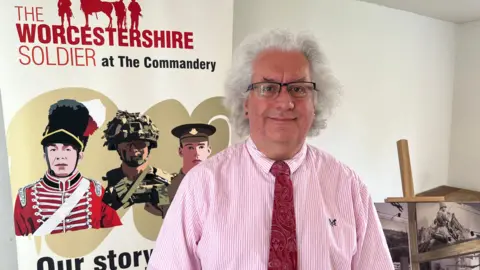 A man stands in front of a sign that reads "The Worcestershire Soldier at The Commandery". He is wearing a pink and white striped shirt with a red patterned tie. He has short grey hair and is wearing a pair of black rimmed glasses. He is smiling.
