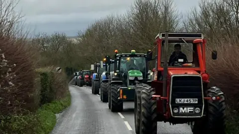 Somerset Tractor Runners A line of six red, green and blue tractors driving along road, which is lined with tall bushes and trees.