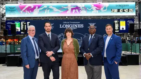 Glasgow 2026 A group of five people, standing in front of a large countdown clock. The group are four men and one woman, Agnes Jones, standing in the centre. She is wearing a tan dress and green cardigan. The four men are all dressed in blue suits. 