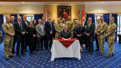 A group of men and women, some in Army uniform, in a room around a small table with a white and red cover. One man is sat at the table holding a pen above the cyber security investment contract