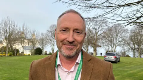A man in a brown blazer and green lanyard smiling in front of garden. 