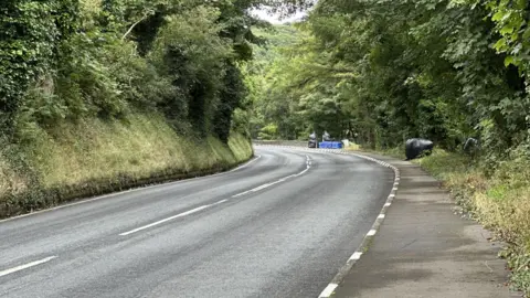 The road sweeping around a left hand bend with a steep grass embankment on the left and a pavement on the right. The are blue and black crash barrier on the right in the distance and trees overhanging on both sides.