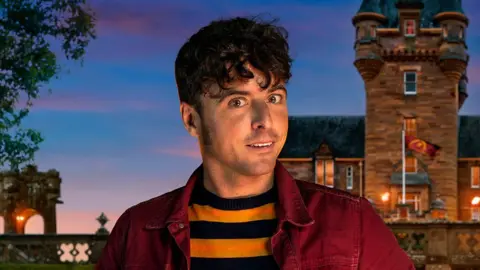 James Baker who has curly black hair, hanging over his forehead, and wearing a red jacket over a yellow and black horizontally striped top. Behind him is a computer generated image of a castle with a turret against a setting sun sky.