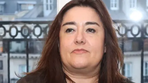 Getty Images Lawyer Francesca Satta sits in front of a window at a news conference. Her full face is framed by long, dark brown hair. She has a determined look about her. 