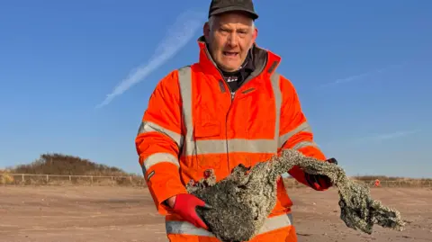 John Byford East Lindsey District Council worker Derrick Whitworth wearing an orange jacket holding a burnt clump of what appears to be plastic pellets on the shoreline at Skegness.