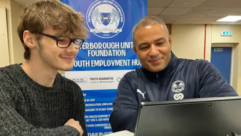 Emma Baugh/BBC Young man wearing a grey jumper and glasses looking at a laptop with an older man with information about the employment hub behind them.