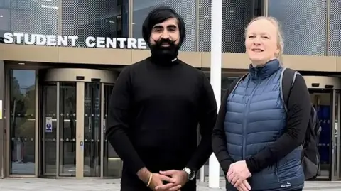 UGC A man with long dark hair and a curled up moustache stands next to a woman with long blond hair. They are outside a university building with a sign that says 'student centre'. 