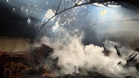 Large plumes of white smoke are coming from a pile of blackened wood, which is on the floor of a barn. Water is being hosed on top. The roof has gaps in it and the steelwork frame is tarnished and black.