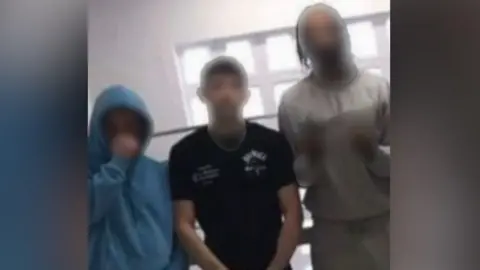 An image showing three men posing behind a stair case and a window. All have their faces blurred out. The man in the middle is wearing all black and posing with his hands down his pants. The man on the left is wearing grey jogging bottoms and a blue hoodie with his hood up. The man on the right is wearing all grey clothes and is making a gesture to the camera with his hands which is also blurred out.