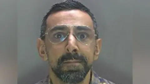 A police photo of Zaheer Abbas showing him looking straight at the camera, with large glasses. He has a beard, short dark hair and is wearing a patterned shirt.