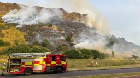 Fire truck and firefighters at foot of a hill which is ablaze