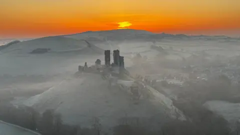 The ruins of Corfe Castle stand on a hill in the Dorset countryside. The surrounding fields are covered in a white frost. The sky is glowing orange as the sun appears over the horizon.