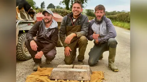 Nick Yallope/Peter Nicolas Three men are crouched down on the ground on what appears to be a road. They are smiling with the two ingots placed on the ground in front of them on a mustard-coloured piece of cloth. An agricultural vehicle with a sheepdog perched on it can be seen behind them.