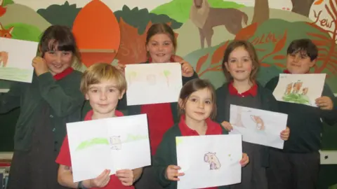 A group of schoolchildren wearing uniform hold up drawings of capybaras. 