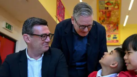 PA Media Andy Burnham (L) has dark hair and spectacles and wears a dark blue suit with white open-necked shirt. Sir Keir Starmer (R) has greying hair, dark spectacles and wears a dark blue suit with an open-necked blue shirt. They talked to two primary school pupils in red sweat shirt and white polo shirt on the PM's trip to Manchester last month.