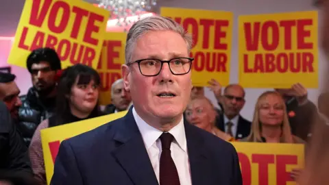 Head and shoulders photo of Keir Starmer looking concerned in front of yellow and red placards with "Vote Labour" printed on them.