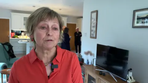 Joe Campbell / BBC A woman in a pink top stood in a living room. She is looking at someone behind the camera.