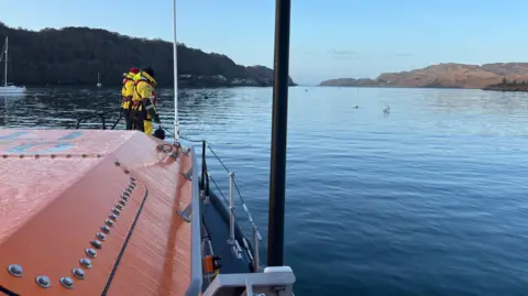 RNLI Oban Lifeboat Station Two RNLI volunteers in yellow survival suits stand at the bow of RNLI Oban's lifeboat. The surface of the sea is calm and there are two headlands in the distance.