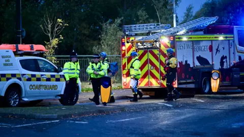 Grimsby News and Pictures Emergency workers gathered infront of a coastguard vehicle and fire engine.  It is still dark but the blue lights light up the surrounding area.