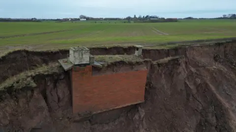 A large brick structure, mainly underground, is sticking out of the side of a clay cliff on a fast-eroding coastline. The bunker has two concrete structures, one a large entrance, the other a squat chimney, above ground. The cliff is dark brown and topped with green grass. Green fields stretch off in the distance towards a village on the horizon. The sky is grey-blue.