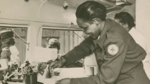 The interior of Clubmobile Massachusetts at a US airbase in East Anglia during World War Two. On the right is a woman in American Red Cross uniform with her black hair pulled back and short. She is leaning over to stir a drink in a mug on a counter and smiling broadly. Behind her is another just glimpsed woman in a white. In front of them are two open windows and American servicemen can be seen drinking from their mugs.