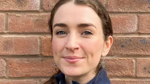 A headshot of a young woman with brown hair tied back. She is standing in front of a brick wall.