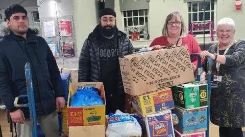 A trolley with boxes of food being handed over by two supermarket staff and Kelly Brown and Trisha Bennett 