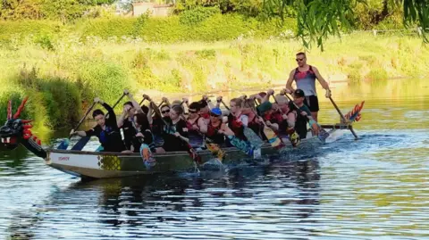 BBC A team of dragon boat racers rowing a boat on a river.