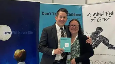 John Adams John Adams is in a black suit with a white shirt, and has his arm around Clare Shaw. She has brown hair and glasses and is wearing a floral dress. He is holding their book which is light blue