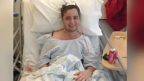 A man in a hospital bed wearing a gown smiles at the camera. He has a drain coming out of his head.