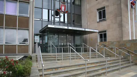 The outside of Shire hall, a stone and glass building with a canopy over the front door topped with a red and white shield. A union jack is flying to the right of the door.