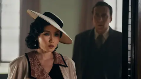 Aaron Epstein/Prime The character of Cat Hardy, a woman wearing a cream coloured hat and top with light red patterns. Behind her is the character of Bill Reilly, wearing a dark coat, tie and white shirt.