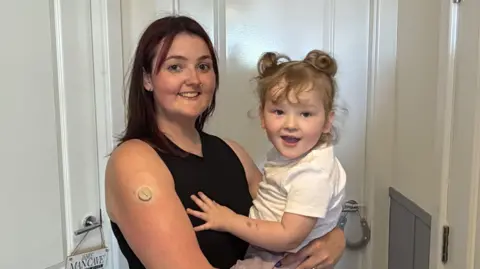 Zoe Mason holding her daughter Izzy in her arms. Mason has mahogany-coloured shoulder length hair and wears a black vest and has a nose ring. Izzy is blond and has curly hair tied in two bunches. She is wearing a white t-shirt. Both are smiling at the camera. 
