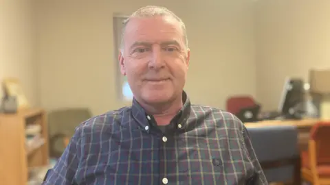 Dean is sitting in a room. He's wearing a checked shirt. The background is blurred. A table abd shelf are in the background. 