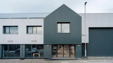 A two storey warehouse building with corrugated panelling. A large service entrance on the right has a dark grey door and light grey panels above it. The reception has glass double doors. Conference spaces on the ground floor to the left also have large windows. A lamp post taller than the warehouse stands just to the right of the entrance.