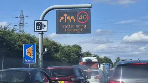 Traffic queues on the motorway. A sign with the text 'Traffic being released' as well as arrows and a temporary speed limit of 40 MPH hangs above.