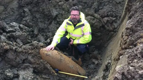 Supplied Mike Sainsbury is crouching behind of a metal item which has been dug out of a muddy patch of ground. The device has a tape measure placed in front of it. He is wearing a yellow high-vis jacket and glasses. He has short, curly black/grey hair and a beard. 