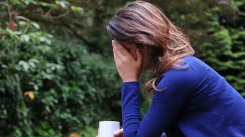 A woman with long brown hair, wearing a long-sleeved blue top, sits down with her hand on her head. She is also holding a mug. 